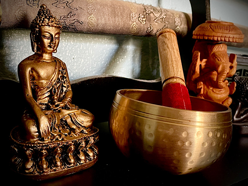 Étagère du salon sur laquelle est posée deux statuettes, une de Bouddha l’autre de Ganesh, ainsi qu’un bol de méditation en metal doré et son marteau.