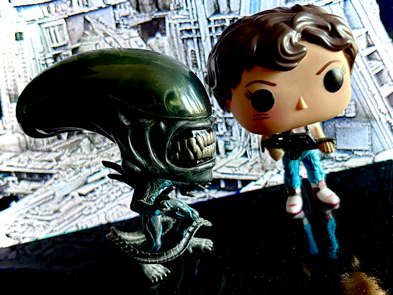 Deux figurines funko pop inspirées de la saga Alien. Elles sont posées sur une surface noire réfléchissante et une photo du Nostromo dans l’espace se trouve en arrière-plan