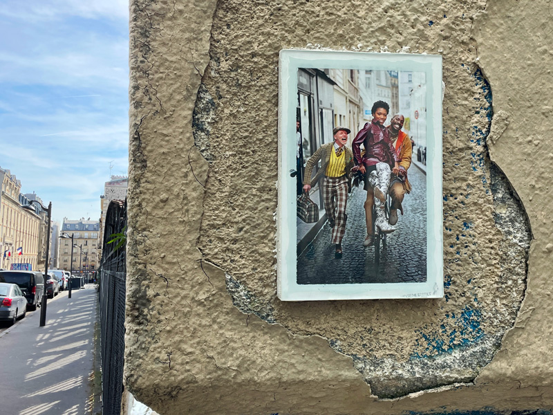 Photo de rue, avec un cadre accroché à un pilonne contenant la photographie d'un trio de personne dont deux sur un vélo.
