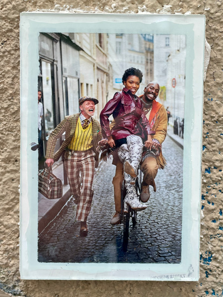 Photographie joyeuse de 2 personnes noires sur un vélo dans une rue pavée. Un homme blanc, plus âgé, en costume a carreaux et casquettes court à côté d'eux.