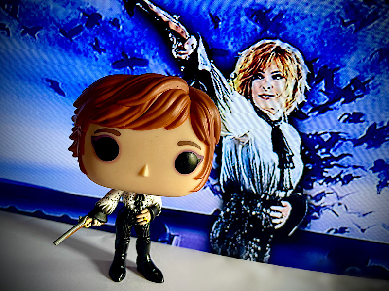 Figurine Funko representant Mylène Farmer dans son costume de scène Libertine lors de la tournée Nevermore. Le fond est une photo de ce moment scénique