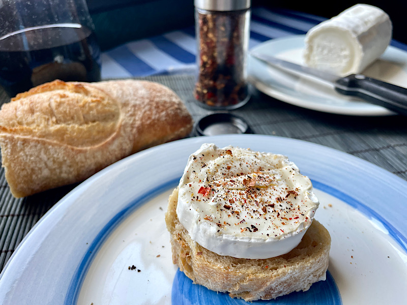 Une assiette avec du fromage de chèvre avec du piment moulu dessus. Un verre de vin, du pain, le moulin à piment et la buche de chèvre en arrière-plan