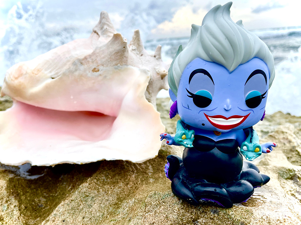 Figurine Ursula Funko sur un rocher avec des vagues qui se brisent derrière elle, la mer et un ciel nuageux où le soleil se lève